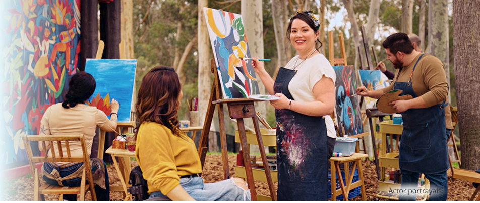 A medium-light skin toned, feminine-presenting person in their thirties painting in an outdoor area. Actor portrayals.