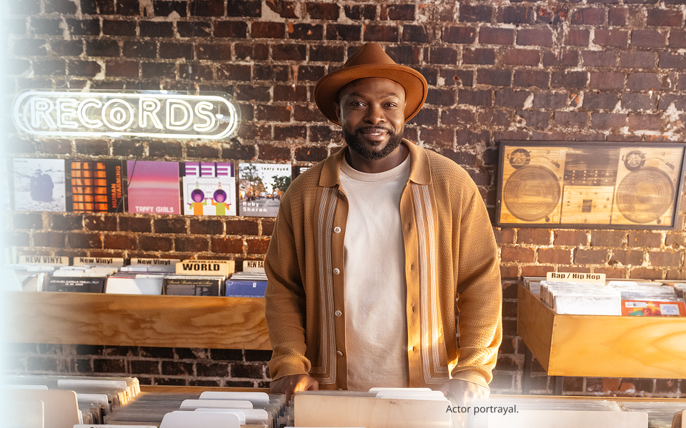 A dark skin toned, masculine-presenting person in their thirties standing in a record store and smiling. Actor portrayal.