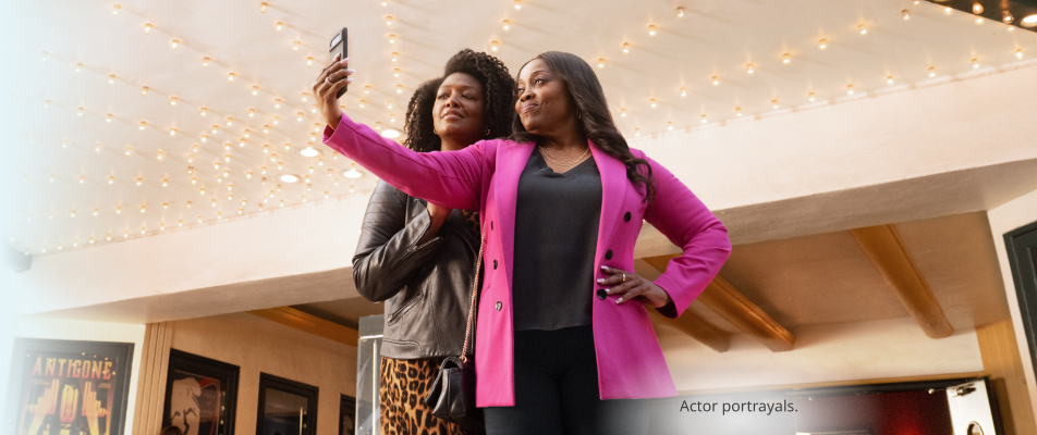 Two dark skin toned, feminine-presenting people standing in front of a movie theater and taking a selfie. Actor portrayals.