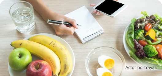 A table filled with healthy food. Light skin toned hands are shown writing and using a phone. Actor portrayal.