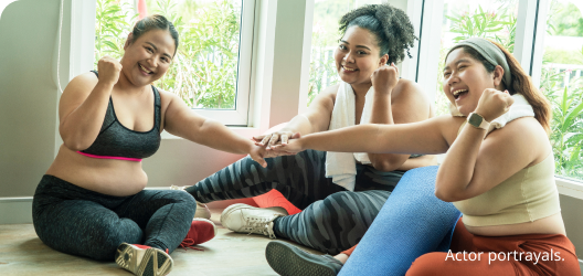 Three feminine-presenting people of various skin tones sitting on the floor of a gym and smiling. Actor portrayals.