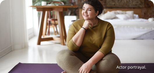 A light skin toned, feminine-presenting person sitting cross-legged on a yoga mat in their bedroom. Actor portrayal.